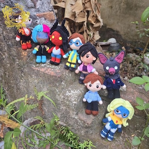 Puede incluir: Una colección de muñecos de ganchillo hechos a mano, cada uno con características y atuendos únicos. Los muñecos están dispuestos sobre una superficie rugosa y texturizada, mostrando una variedad de colores y diseños. Los muñecos parecen estar inspirados en personajes de dibujos animados o películas.