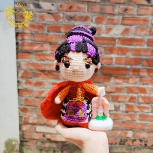 May include: Crocheted doll with purple hair, a red cape, and a purple and orange dress. The doll is holding a white and black crocheted broom.