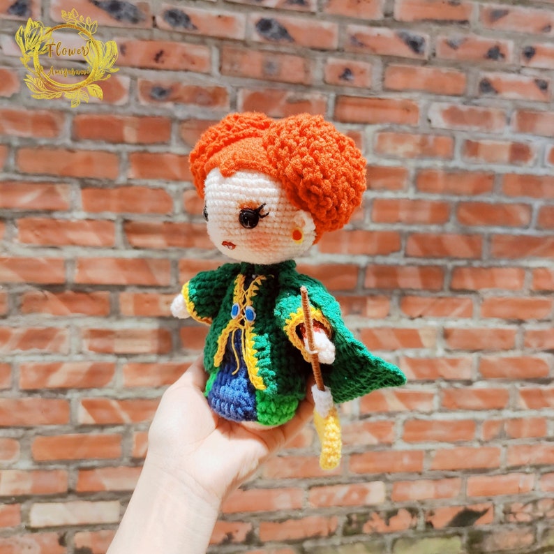 May include: Crocheted doll with red hair in two buns, wearing a green and blue dress with a yellow sash and holding a brown wand.
