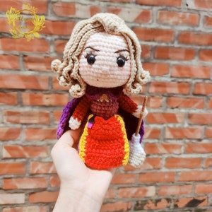 May include: Crocheted doll with blonde hair, wearing a red and purple dress with a yellow trim, holding a broom. The doll has a cartoonish face with large eyes.