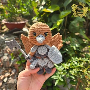 以下が含まれることがあります： 手作りのかぎ針編みフクロウの置物。茶色の頭と羽、灰色の鎧と盾が特徴です。フクロウは小さな灰色の斧を持っています。コレクターや装飾品として最適な、ユニークな手作りのアイテムです。