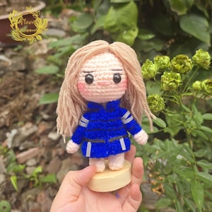 May include: A handmade crochet doll with blonde hair and a blue jacket with white stripes. The doll has a light skin tone, rosy cheeks, and is standing on a small wooden base. The doll is a charming, handcrafted item.