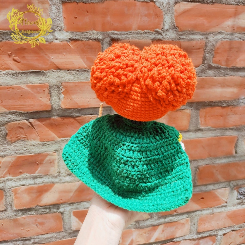 May include: A crocheted doll with a green hat and orange hair. The doll is handmade and has a whimsical look.