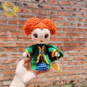 May include: A crocheted doll with red hair in two buns, wearing a green and yellow robe and holding a broom. The doll has a white face with black eyes and a red mouth.