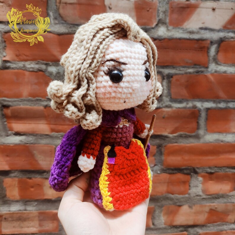May include: Crocheted doll with blonde hair, wearing a purple and red dress with a yellow trim. The doll is holding a stick in its right hand.
