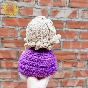 May include: A crocheted doll with a purple dress and a beige crocheted wig. The doll is being held by a hand.