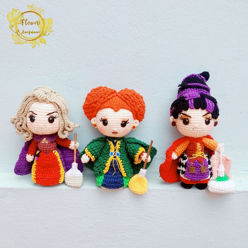 May include: Three crocheted witch dolls with different colored outfits and accessories. The doll on the left is wearing a red dress with a purple cape and holding a broom. The doll in the middle is wearing a green dress with a yellow cape and holding a broom. The doll on the right is wearing a purple dress with a red cape and holding a bag.