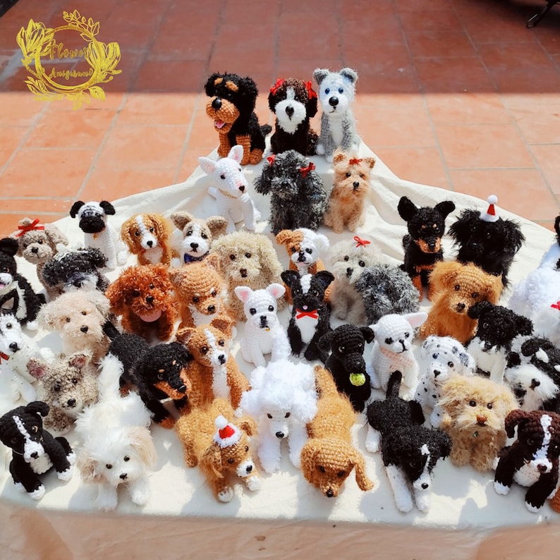May include: A collection of crocheted dog toys in various breeds and colors, including black, white, brown, and gray. The toys are arranged on a white surface.