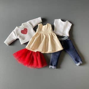 May include: A collection of doll clothes including a white sweatshirt with a red glitter heart, a cream-colored polka dot dress, a red tulle skirt, a white t-shirt, and blue denim jeans. The clothes are arranged on a gray surface.