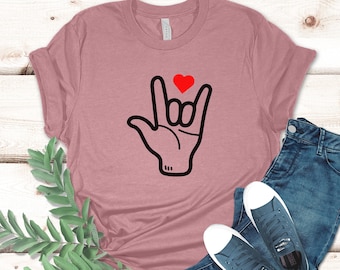 May include: Dusty rose t-shirt with a black hand sign and a red heart. The hand sign is a symbol of love in American Sign Language. The shirt is paired with blue jeans and white-soled sneakers.
