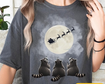 May include: Dark gray t-shirt featuring a whimsical Christmas design. The graphic shows three raccoons looking up at Santa's sleigh flying in front of a full moon. The word "PEPPER" is printed on a yellow rectangle.
