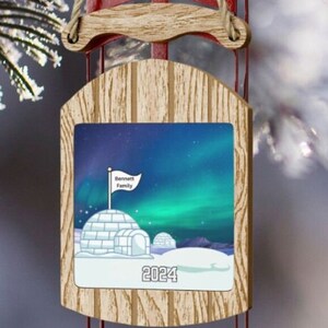 May include: A wooden sled Christmas ornament with a white flag that says "Bennett Family" and a scene of two igloos in front of a green and blue aurora borealis. The year 2024 is printed on the snow.