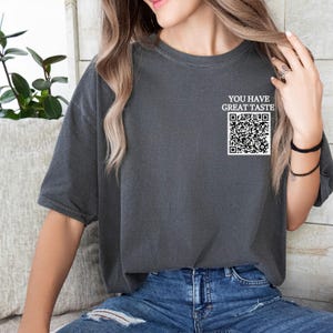 May include: A dark gray t-shirt with the text "YOU HAVE GREAT TASTE" and a black and white QR code printed on the front.