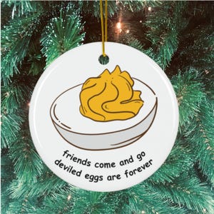 May include: White ceramic Christmas ornament featuring a deviled egg illustration with a yellow filling. The ornament has the text "friends come and go, deviled eggs are forever" in black script. A gold string is attached for hanging.