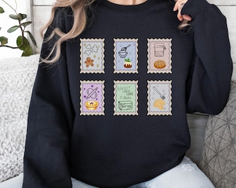 May include: Black sweatshirt featuring six postage stamp-style graphics with baking-themed illustrations. Designs include a gingerbread man, mixing bowl, jar of bread, pie, butter, and a whisk. The text "Baked with Love & Butter" is also featured.