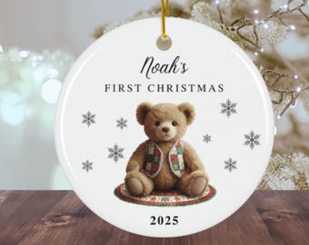 May include: A white ceramic Christmas ornament with a teddy bear illustration. The bear wears a vest with a colorful pattern and sits on a patterned rug. The ornament reads "Noah's FIRST CHRISTMAS 2025" with snowflake accents and a gold string.