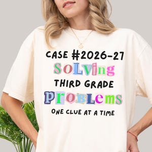 May include: A cream-colored short-sleeve t-shirt featuring the black text CASE #2026-27, THIRD GRADE, and ONE CLUE AT A TIME. The words SOLVING and PROBLEMS are displayed in a colorful, multi-patterned ransom note style font with varied letters.