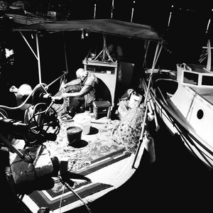 Könnte beinhalten: Ein Schwarzweißfoto eines Fischers auf einem Boot bei Nacht, der ein Netz einzieht. Das Boot wird von einem einzigen Licht beleuchtet, und der Fischer trägt einen Hut und eine Jacke. Das Netz ist hoch auf dem Deck des Bootes gestapelt.