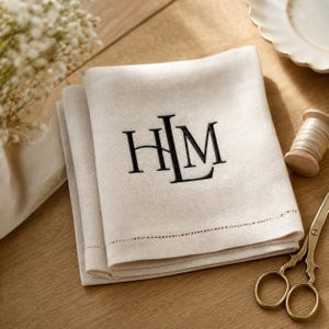 May include: A folded beige linen napkin featuring a black embroidered monogram with the letters H, L, and M. The napkin has a decorative hemstitch border detail along the bottom edge, showcasing a clean, woven fabric texture and neutral tone.