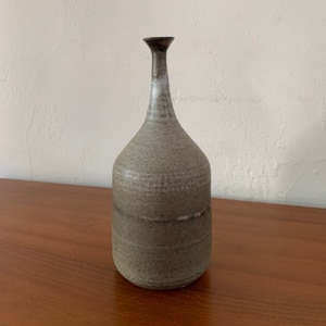 May include: A handcrafted ceramic vase with a slender neck and a bulbous body. The vase has a speckled, earthy gray-green color with a dark brown rim. The vase is approximately 8 inches tall, making it a decorative piece for any room.