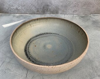 May include: A large, round, ceramic bowl with a speckled, light brown glaze. The bowl has a light blue interior with a dark gray ring around the center.