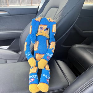 May include: A handmade stuffed toy shaped like a rabbit, crafted from blue fabric featuring a repeating pattern of orange and white Kraft Singles cheese slices. The toy has long ears, yellow fabric feet, and two mismatched button eyes in purple and black.