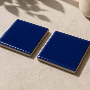 Puede incluir: Dos posavasos cuadrados de cerámica azul marino con acabado brillante. Los posavasos tienen un borde beige claro y están colocados sobre una superficie neutra. Un artículo de decoración para el hogar.