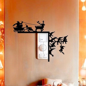 Puede incluir: Una calcomanía de pared en silueta negra de Santa Claus en un trineo tirado por renos con duendes colgados del trineo. La calcomanía se coloca en una pared junto a un interruptor de luz.