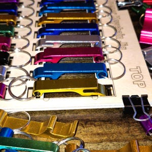 May include: A close-up of a set of colorful bottle openers on keychains. The openers are arranged in a row on a wooden board with the word "TOP" printed on it. The openers are in various colors, including gold, silver, blue, pink, and green.