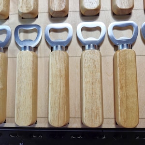 May include: A collection of bottle openers with wooden handles and silver-colored metal tops. The openers are arranged in a wooden display case, showcasing their natural wood grain and simple design. The bottle openers are designed for opening bottles.