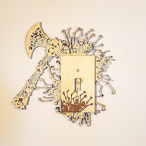 May include: A light switch cover with a wood-carved design of skeletons and bones. The design is intricate and detailed, with the skeletons appearing to be reaching out from the sides of the switch cover.