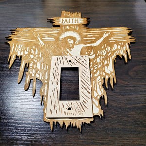 Op de afbeelding: Een houten lichtschakelaar-afdekplaat met een uitgesneden ontwerp van een persoon op een kruis. Het ontwerp is in tinten bruin en wit. Het woord "FAITH" staat op het kruis.