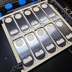 May include: A collection of ten stainless steel bottle openers, each with a circular handle and a bottle cap opener. They are arranged in a wooden tray with individual cutouts. The openers have a brushed metal finish.