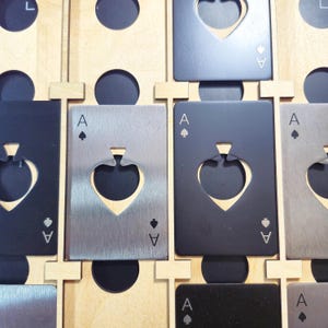 Op de afbeelding: Close-up van verschillende metalen Ace of Spades flesopeners in verschillende tinten grijs en zwart. Elke opener is in een houten display geplaatst met een schoppenvormige uitsparing. Het Ace of Spades-symbool is op elke opener geëtst.
