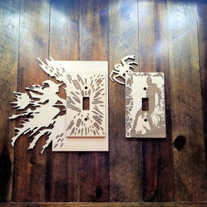 Könnte beinhalten: Lichtschalterabdeckungen aus Holz mit lasergeschnittenen Designs. Eines zeigt eine Hexe auf einem Besen, ein anderes ein Burst-Muster und das dritte eine zombieartige Figur. Eine Holzspinne ist ebenfalls enthalten. Die Abdeckungen sind an einer Holzwand befestigt.