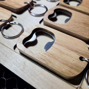 Peut inclure: Porte-clés décapsuleurs en bois avec un ouvre-bouteille en métal argenté. Les porte-clés sont en bois de couleur claire et ont une forme rectangulaire avec une découpe pour l'ouvre-bouteille. Les porte-clés sont attachés à des anneaux argentés.