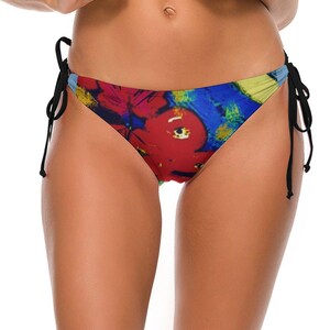May include: A bikini bottom featuring a vibrant abstract print with bold red, deep blue, and yellow brushstroke patterns. The garment includes thin black side ties that are knotted at each hip, providing an adjustable fit for this swimwear piece.