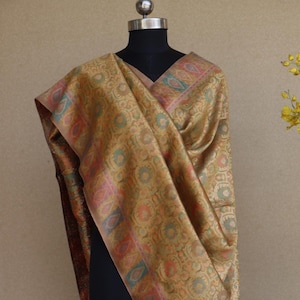 Peut inclure: Un châle doré avec un motif floral complexe et une bordure décorative aux tons rose, bleu et orange. Le châle est drapé sur un mannequin noir, mettant en valeur son design et sa texture. Les bords sont frangés.
