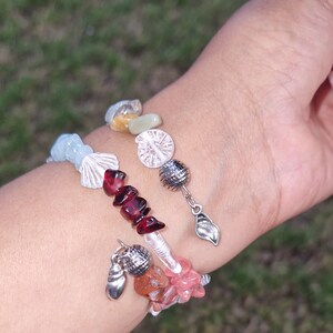 May include: Two bracelets with various colored stones and silver charms. The top bracelet has light blue, white, red, and clear stones with a silver charm of a leaf. The bottom bracelet has orange, pink, and clear stones with a silver charm of a seashell.