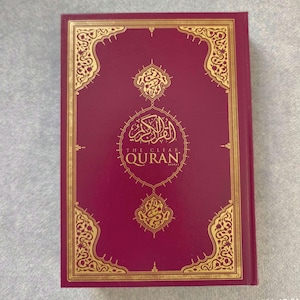 以下が含まれることがあります： 金色の装飾と「THE CLEAR QURAN」のタイトルが金色の文字で書かれた、バーガンディ色の本。表紙には、華やかな金色の模様とアラビア語のカリグラフィーが施されています。本は長方形で、新品のようです。