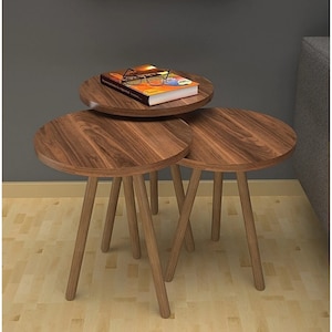 May include: Three round wooden nesting tables with a natural wood finish and three legs each. The top table has a book and glasses on it.
