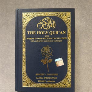 May include: A navy blue book titled "THE HOLY QUR'AN" with gold ornate detailing. The cover includes Arabic script and the text "WORD BY WORD ENGLISH TRANSLATION". The book is published by RAYHA PUBLICATION in Turkey.
