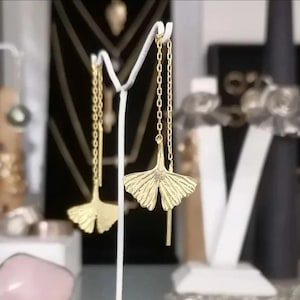 May include: Gold tone earrings with a ginkgo leaf design. The earrings are on a white display stand.
