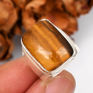 May include: A silver ring featuring a large, rectangular tiger's eye stone. The stone displays warm brown and gold tones with natural striations. The ring has a simple, modern design, showcasing the gemstone's natural beauty. The ring is held by a person's fingers.