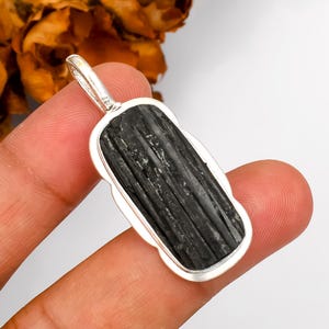 Peut inclure: Un pendentif rectangulaire avec une pierre noire texturée sertie dans un cadre en métal argenté. Le pendentif a une boucle en haut pour une chaîne. La pierre a un aspect naturel et strié.