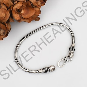 Puede incluir: Una pulsera de cadena de serpiente de plata con un diseño de metal trenzado y texturizado. La pulsera incluye dos tapas cilíndricas decorativas con detalles intrincados y un mecanismo de cierre de gancho y ojo seguro.