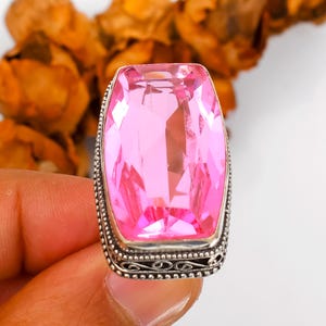 May include: A large, rectangular pink gemstone ring with a silver-colored band. The gemstone is faceted and catches the light, showcasing its vibrant color. The band features an ornate, detailed design.