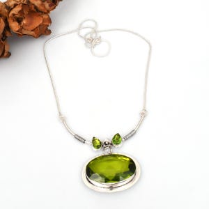 May include: A silver necklace featuring a large, oval-shaped, lime-green gemstone pendant. Two smaller green gemstones are set above the pendant. The necklace has a delicate silver chain and a curved bar with wire detailing.