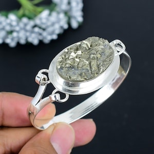 Puede incluir: Una pulsera de plata con una piedra de pirita ovalada. La piedra de pirita es de color gris metálico texturizado, engastada en un bisel de plata. La pulsera tiene un diseño articulado con cierre de broche.