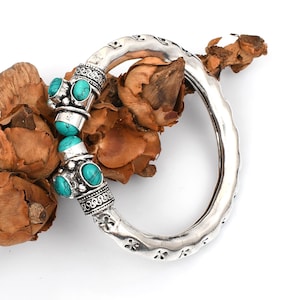 May include: A silver-colored bangle bracelet with turquoise-colored stones. The bracelet has an oval shape with decorative detailing and is adorned with three turquoise stones. The bracelet is displayed on a bed of dried, brown plant material.